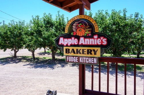 Sign for Apple Annie's Bakery and Fudge Kitchen, surrounded by apple trees in a sunny outdoor setting.