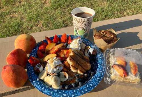 A colorful breakfast spread with pancakes, fruit, coffee, and a muffin on a picnic table outdoors.