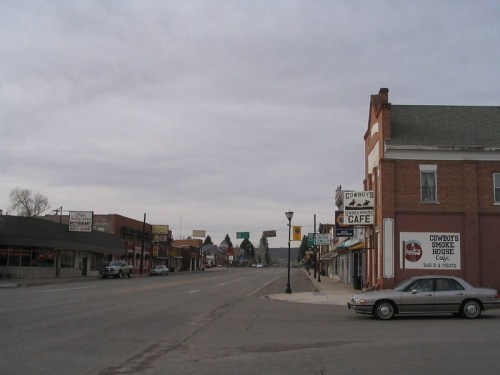 A quiet street scene in a small town, featuring shops, a café, and a parked car under a cloudy sky.