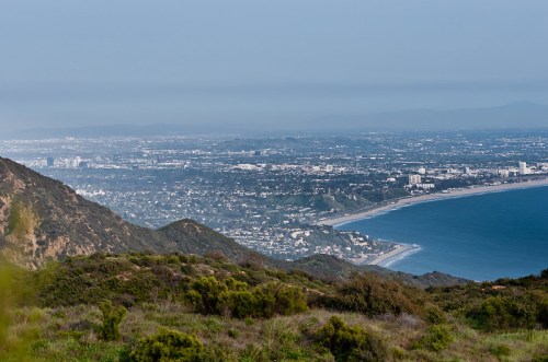 A panoramic view of a coastal city with mountains in the foreground and the ocean in the background under a hazy sky.