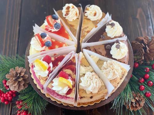 A circular platter of assorted cheesecake slices, decorated with fruit and whipped cream, surrounded by pinecones and greenery.