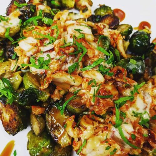 A plate of roasted Brussels sprouts topped with shredded chicken, cheese, and a drizzle of sauce, garnished with herbs.