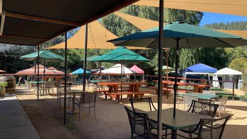 Outdoor seating area with tables and umbrellas, surrounded by colorful tents and trees in a sunny setting.
