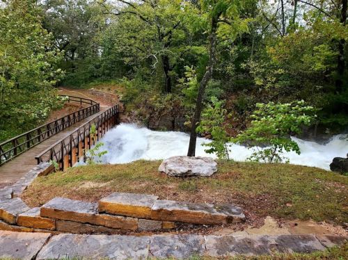 A wooden bridge crosses a rushing river, surrounded by lush green trees and rocky terrain.