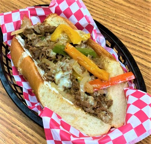 A loaded sandwich with sliced beef, sautéed onions, and colorful bell peppers in a soft hoagie roll, served in a basket.
