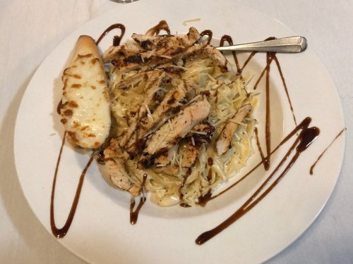 Grilled chicken strips on a bed of pasta, drizzled with sauce, served with a cheesy breadstick on the side.