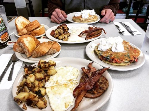 A table set with plates of pancakes, eggs, bacon, and toast, with a person’s hands visible in the background.