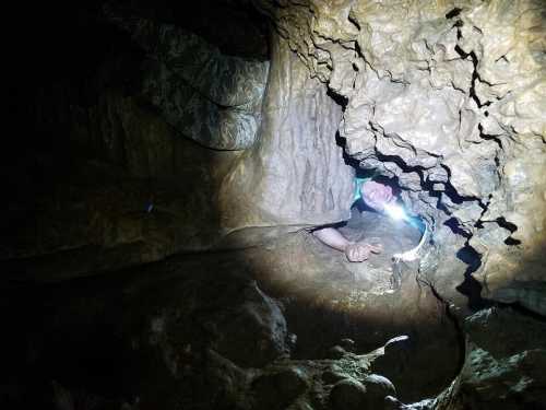 A person with a flashlight is partially emerging from a narrow cave opening, surrounded by rocky formations.