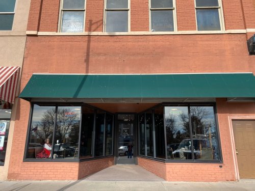 A storefront with a green awning and large windows, featuring a brick facade and a door on the right side.