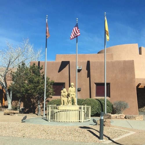 A statue of two figures stands in a courtyard, surrounded by flags and desert landscaping under a clear blue sky.