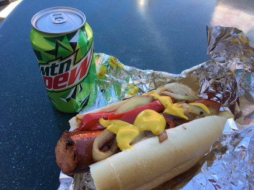A hot dog topped with onions, tomatoes, and mustard, next to a can of Mountain Dew on a table.