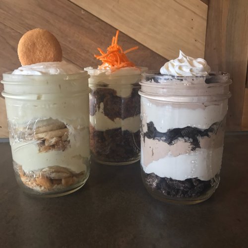 Three layered desserts in mason jars: vanilla with cookies, carrot cake, and chocolate with Oreos, topped with whipped cream.