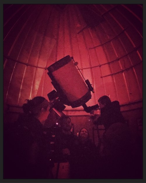 A group of people inside a dimly lit observatory, gathered around a large telescope.