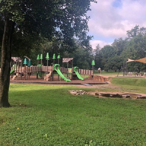 A playground with green slides and wooden structures, surrounded by trees and grassy areas under a cloudy sky.