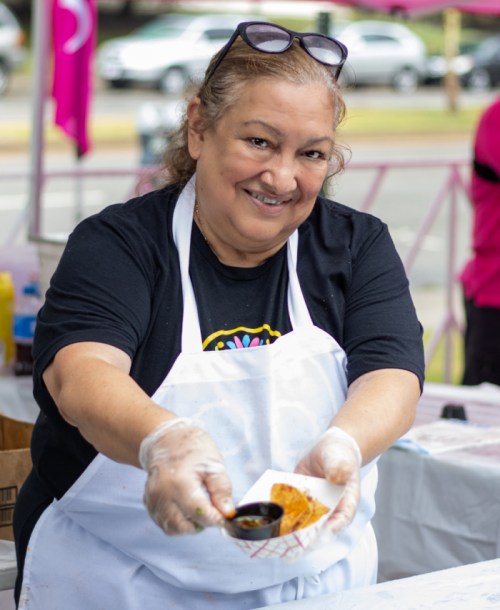 A smiling woman in an apron holds a small dish of food with sauce, standing at a food stall.