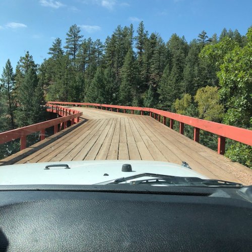 A wooden bridge with red railings winding through a forest, viewed from the front of a vehicle.