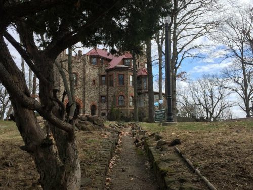 A stone castle with a red roof, surrounded by trees and a grassy path leading up to it under a cloudy sky.