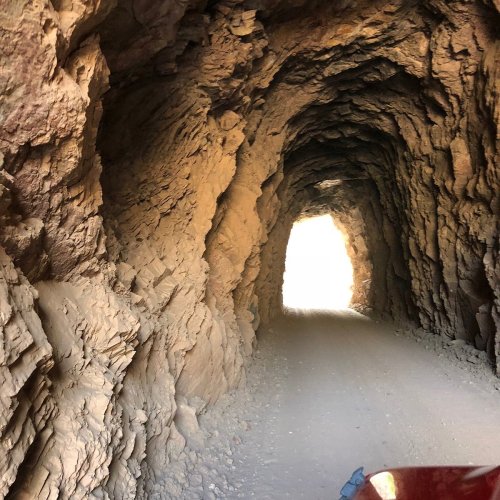 A rocky tunnel with a dirt road leading to a bright light at the end. The walls are rugged and textured.