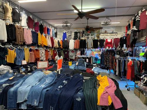 A clothing store interior filled with racks of colorful tops and jeans, with a ceiling fan overhead.