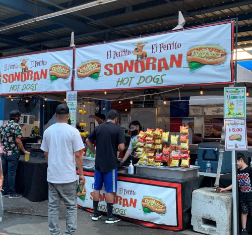 A hot dog stand named "El Perrito Sonoran" with customers and snacks displayed in a market setting.