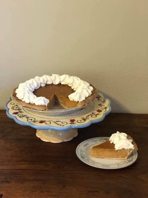A pumpkin pie with whipped cream on a decorative cake stand, with a slice served on a plate beside it.