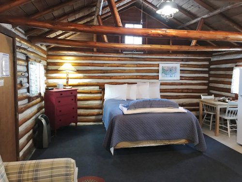 Cozy log cabin interior featuring a bed, red dresser, lamp, and dining area with rustic wooden walls.