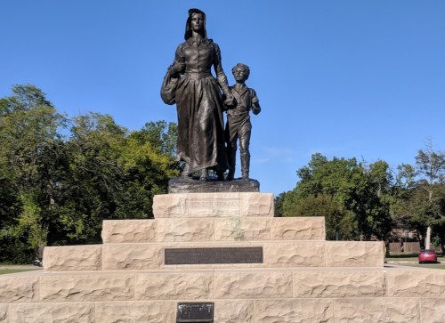 A bronze statue of a woman holding a child's hand, standing on a stone pedestal in a park setting.