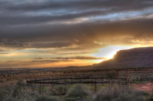 A scenic landscape at sunset, featuring a mountain silhouette, cloudy sky, and a wooden fence in the foreground.