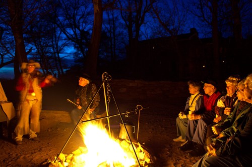 A group of people sits around a campfire at dusk, enjoying a performance by a musician in a wooded area.