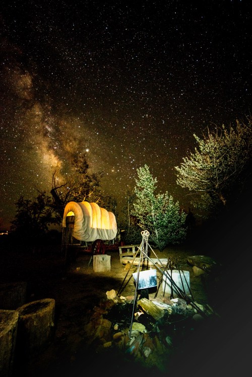 A covered wagon under a starry night sky, with the Milky Way visible and a campfire setup nearby.