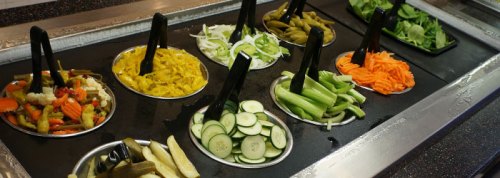 A salad bar featuring various fresh vegetables, including carrots, cucumbers, and leafy greens, with serving tongs.