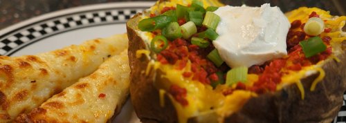 A loaded baked potato topped with cheese, bacon bits, green onions, and sour cream, served with cheesy breadsticks.