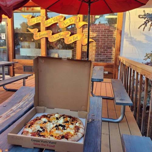 A pizza box sits on a picnic table outside a restaurant with red umbrellas and a welcoming sign in the background.