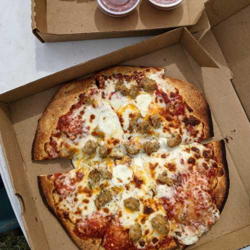 A pizza with melted cheese, pepperoni, and sausage, served in a cardboard box alongside dipping sauces.