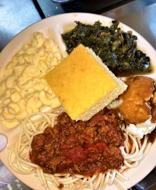 A plate of food featuring macaroni and cheese, greens, cornbread, spaghetti with sauce, and a serving of mashed potatoes.
