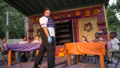 A performer in a costume entertains a group of children seated at colorful tables in a festive outdoor setting.
