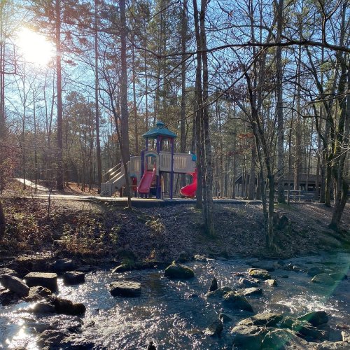 A playground with slides and climbing structures surrounded by trees, next to a stream on a sunny day.