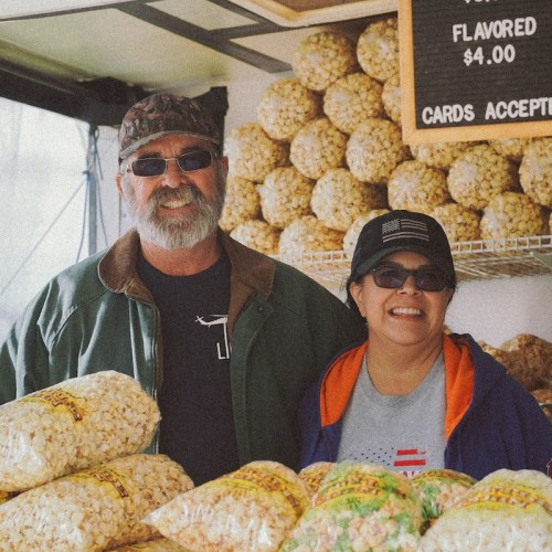 A man and woman smile behind a popcorn stand filled with bags of popcorn. Signs indicate prices and payment options.