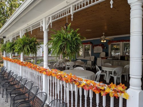 A cozy outdoor dining area with white tables and chairs, decorated with autumn leaves and hanging ferns.