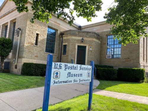 Exterior of the U.S. Postal Service Museum, featuring a stone building and a sign in front. Green grass and bushes surround it.