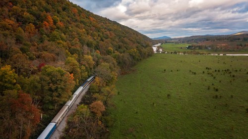 A train travels along a scenic route through colorful autumn trees and a lush green valley under a cloudy sky.