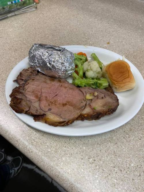 A plate with sliced roast beef, a baked potato in foil, mixed vegetables, and a dinner roll on a countertop.