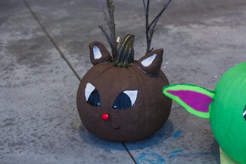 A painted brown pumpkin with antlers, ears, and a red nose, resembling a reindeer, sits on a concrete surface.