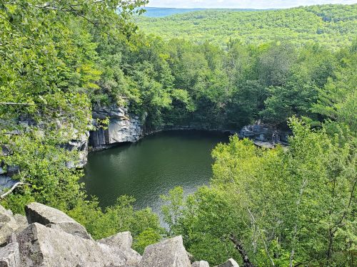 A serene, green landscape surrounds a dark, still pond nestled among rocky cliffs and trees.