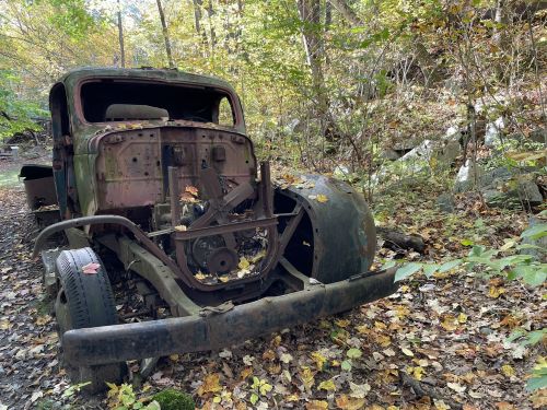 An old, rusted truck sits abandoned in a forest, surrounded by colorful autumn leaves and trees.