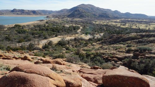 A scenic view of rolling hills and a lake, with rocky terrain in the foreground and sparse trees in the landscape.
