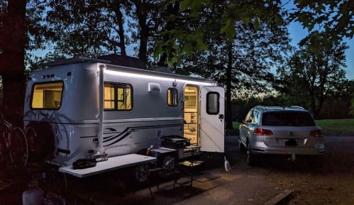 A camper trailer with lights on, parked beside a car, surrounded by trees at dusk. A table is set up outside.
