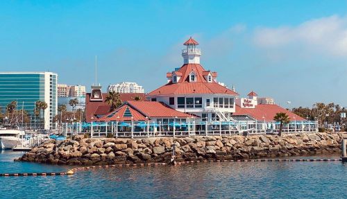 A waterfront building with a red roof and white trim, surrounded by water and palm trees, under a clear blue sky.