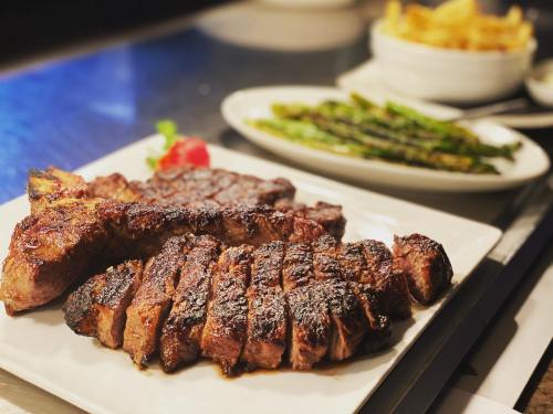 A plate of grilled steak with a garnish, accompanied by asparagus and a bowl of fries in the background.