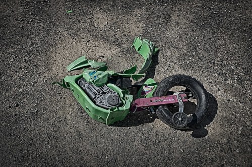 A broken green toy motorcycle lies on a gravel surface, with scattered pieces and a detached wheel.